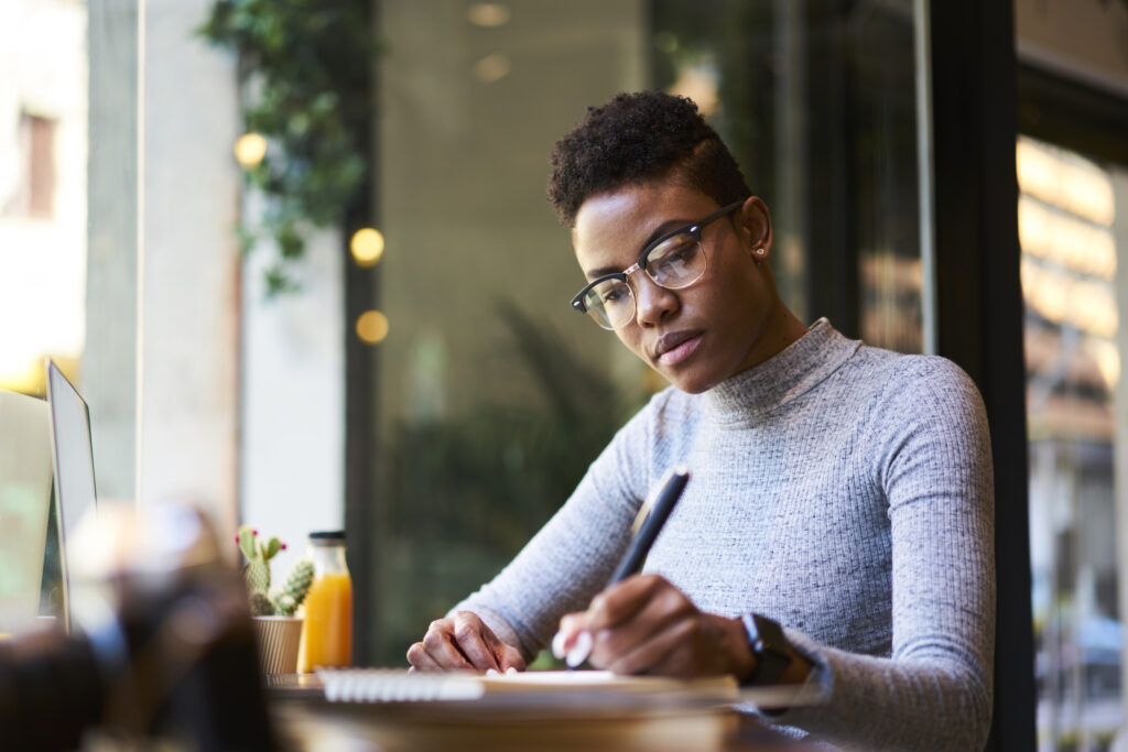 A young African-American woman wearing glasses sitting at a desk writing in a notepad.