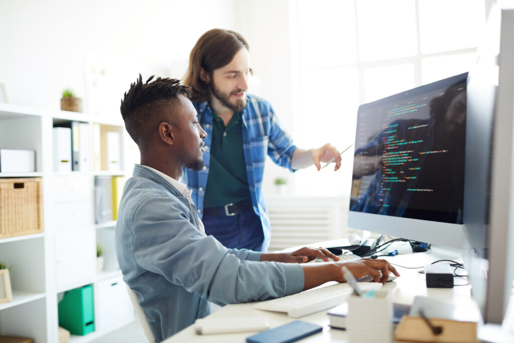 A young African-American man seated at a desk looking at a computer screen with his hands on a keyboard and mouse. Standing to his left is a young white man pointing to the computer screen.